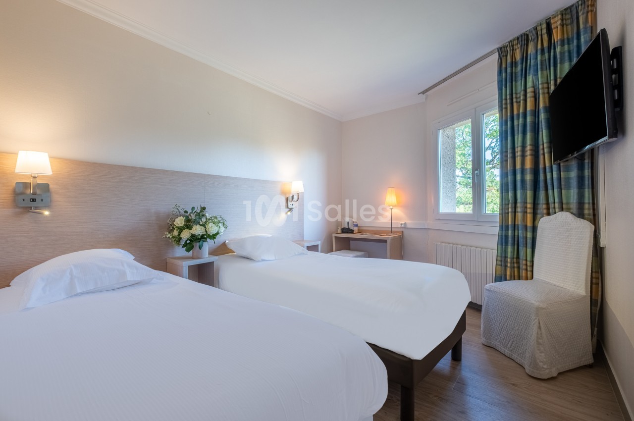 Chambre lumineuse avec deux lits simples, une chaise, une télévision murale et une fenêtre avec rideaux colorés.