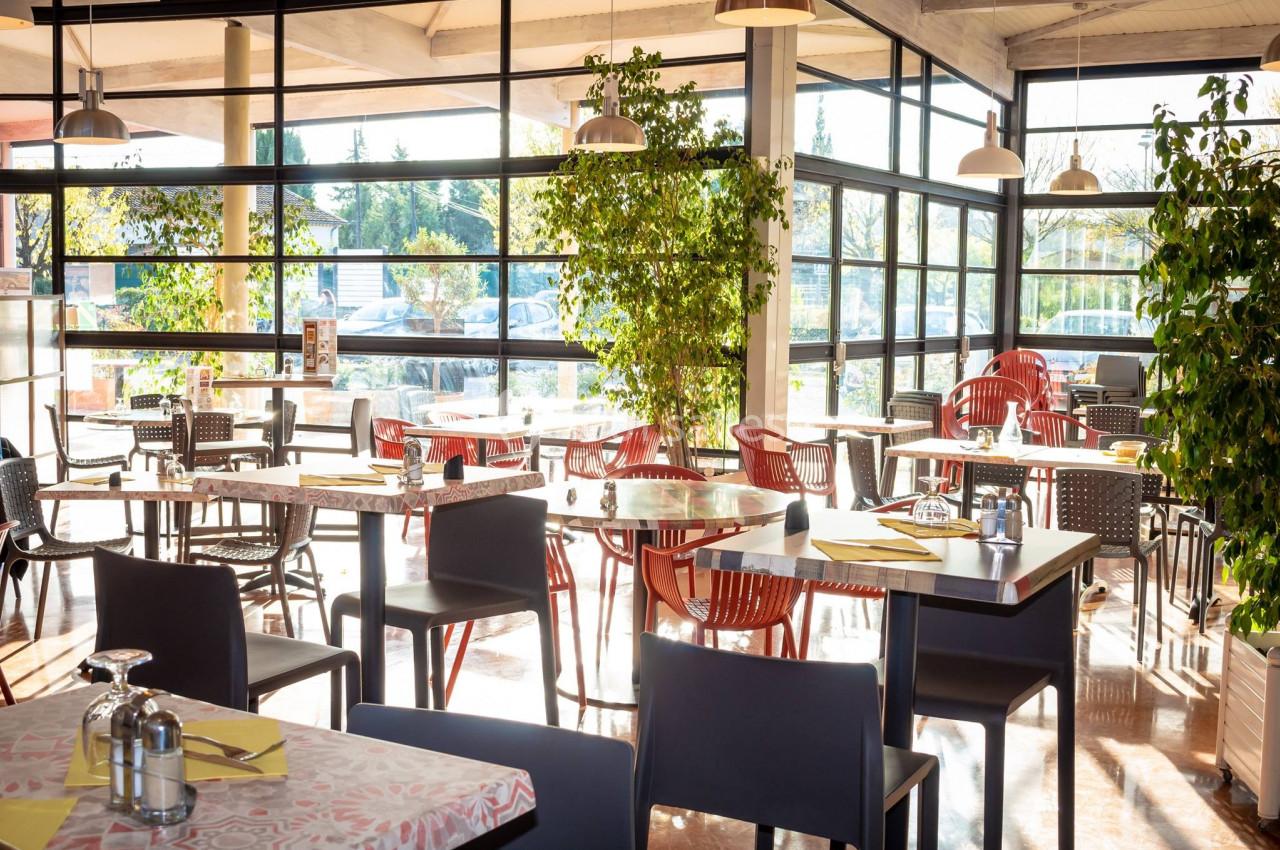 Salle de restaurant lumineuse avec des tables dressées, des chaises colorées et des plantes près de grandes baies vitrées.