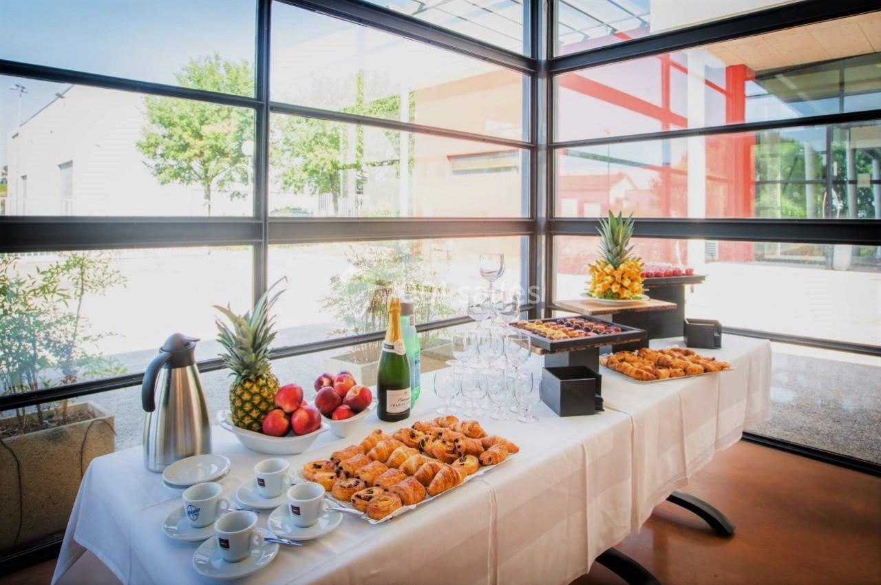 Table dressée avec viennoiseries, fruits, boissons et verres dans une salle lumineuse avec vue sur l'extérieur.