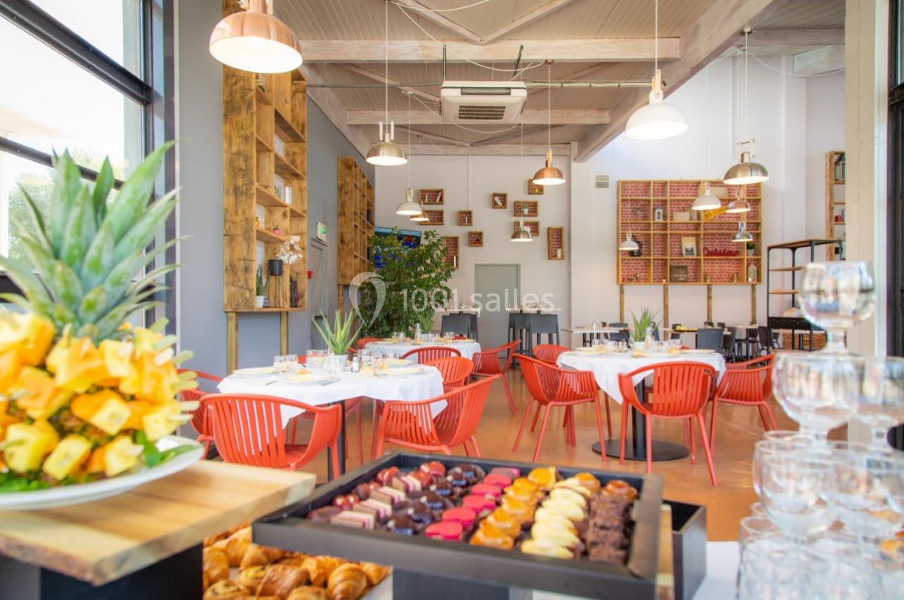 Salle de restaurant lumineuse avec tables dressées, chaises rouges et buffet de pâtisseries et fruits au premier plan.
