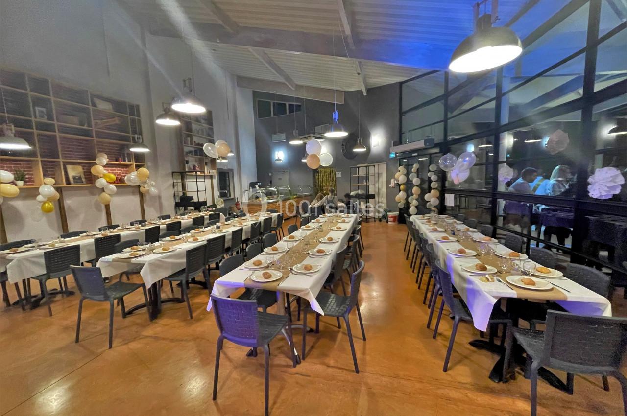 Salle décorée pour un événement, avec deux longues tables dressées, chaises noires et guirlandes suspendues.