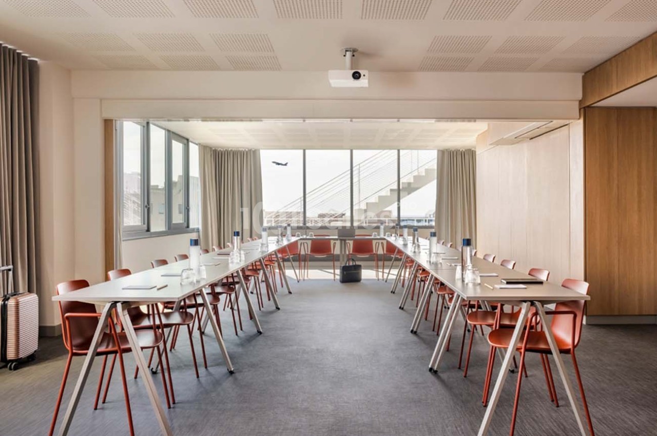 Salle de réunion lumineuse avec tables en U, chaises rouges, grandes fenêtres et projecteur au plafond.