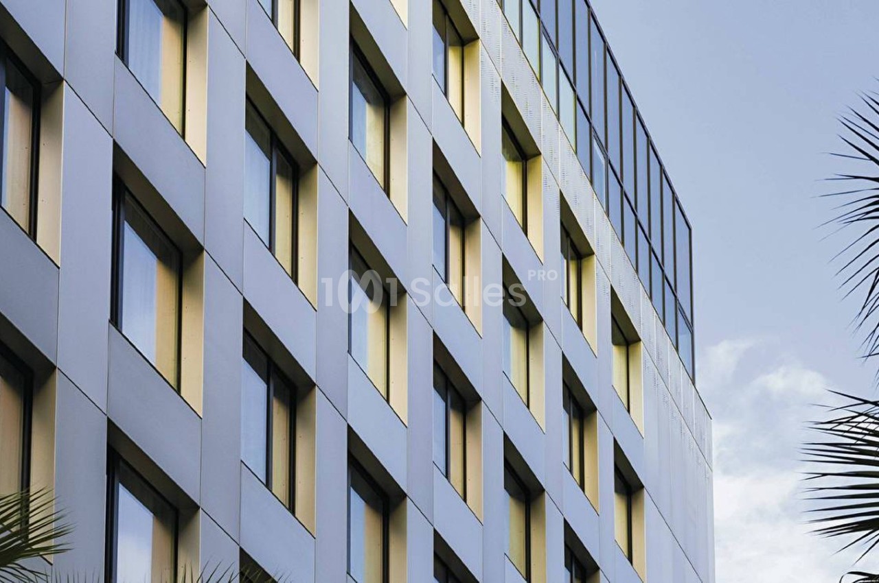 Façade d'un immeuble moderne avec des fenêtres alignées, entourée de palmiers sous un ciel partiellement dégagé.