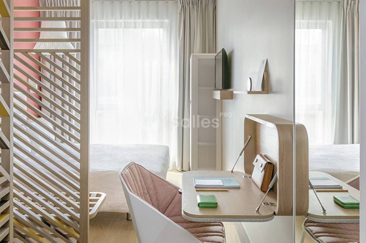 Bureau compact en bois clair avec chaise rose, situé dans une chambre lumineuse séparée par une cloison ajourée.