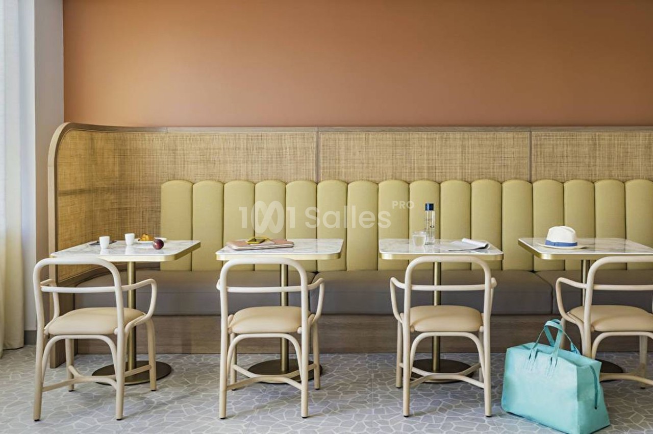 Salle de restaurant avec banquette jaune, tables en marbre, chaises en bois clair et sac turquoise posé au sol.