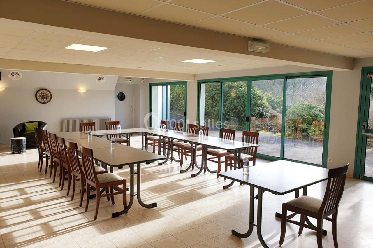 Salle lumineuse avec tables et chaises disposées en U, grandes baies vitrées donnant sur un jardin.