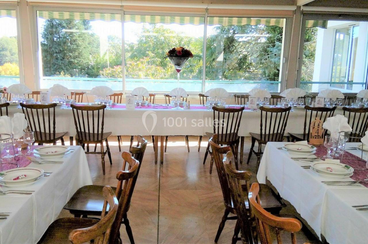 Salle de réception lumineuse avec tables dressées, nappes blanches, chaises en bois et vue sur un jardin à travers de…