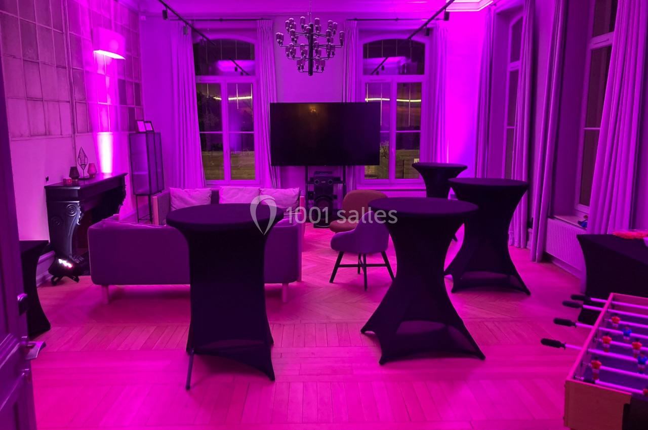 Location salle Lille (Nord) - Les Salons de l'Urban #6 Salle lumineuse avec éclairage rose, tables hautes noires, canapé, télévision murale et baby-foot au premier plan.