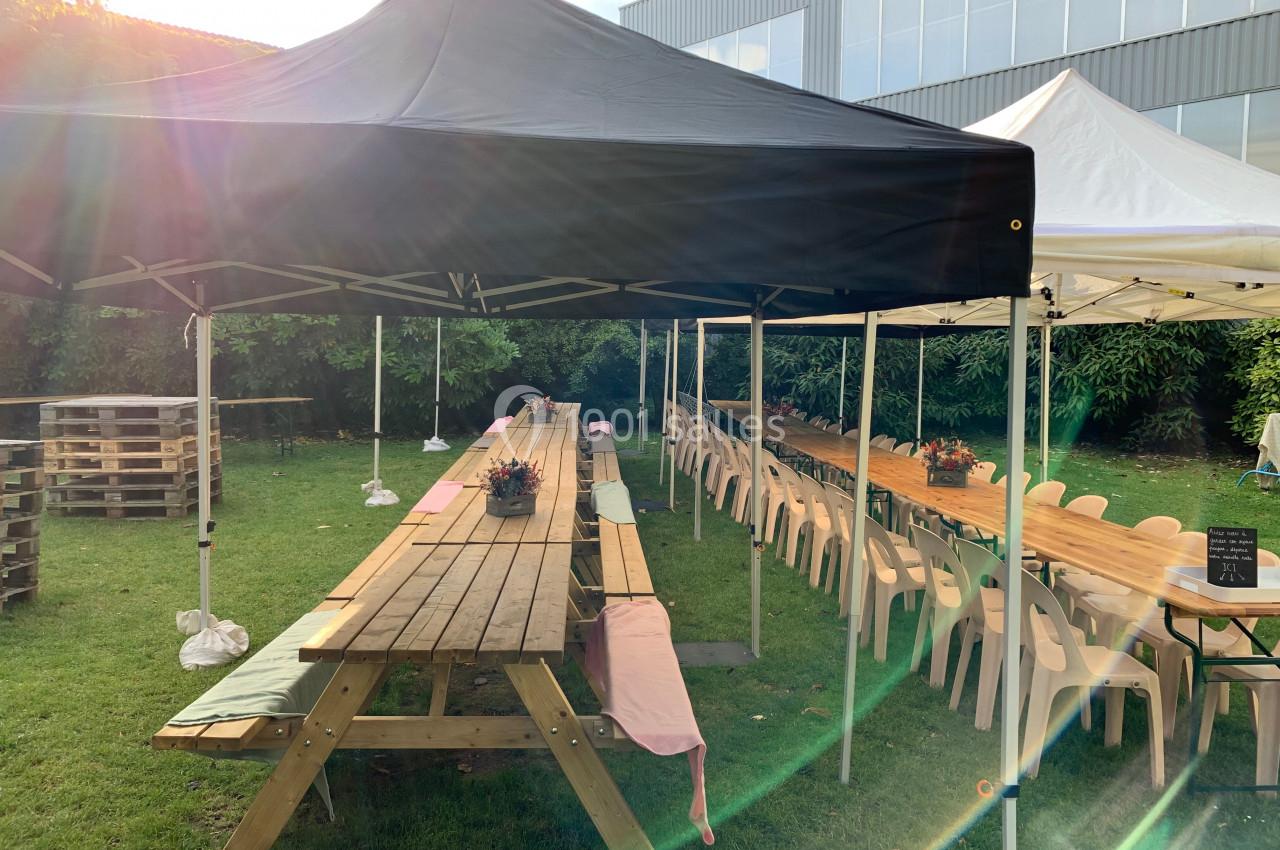 Location salle Lille (Nord) - Les Salons de l'Urban #12 Tables en bois alignées sous des tentes noires et blanches dans un jardin, entourées de chaises et de verdure.
