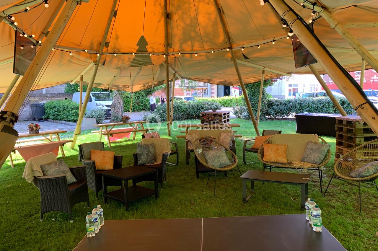 Location salle Lille (Nord) - Les Salons de l'Urban #27 Espace extérieur aménagé sous une tente avec tables, fauteuils, guirlandes lumineuses et verdure en arrière-plan.