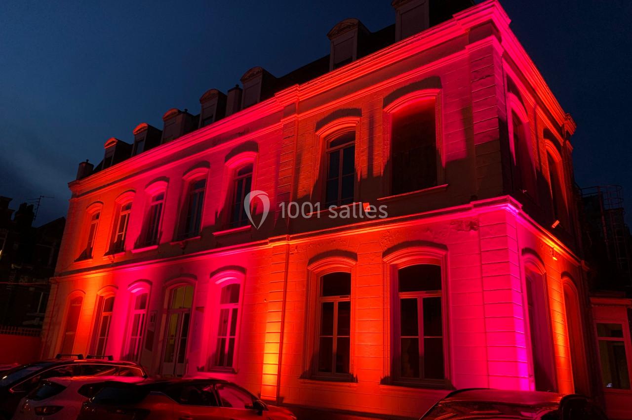 Location salle Lille (Nord) - Les Salons de l'Urban #11 Façade d'un bâtiment éclairée par des lumières roses et oranges, avec des voitures garées au premier plan.