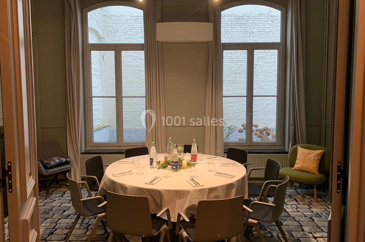 Location salle Lille (Nord) - Les Salons de l'Urban #34 Salle lumineuse avec une table ronde dressée, entourée de chaises, devant de grandes fenêtres donnant sur un mur en briques…