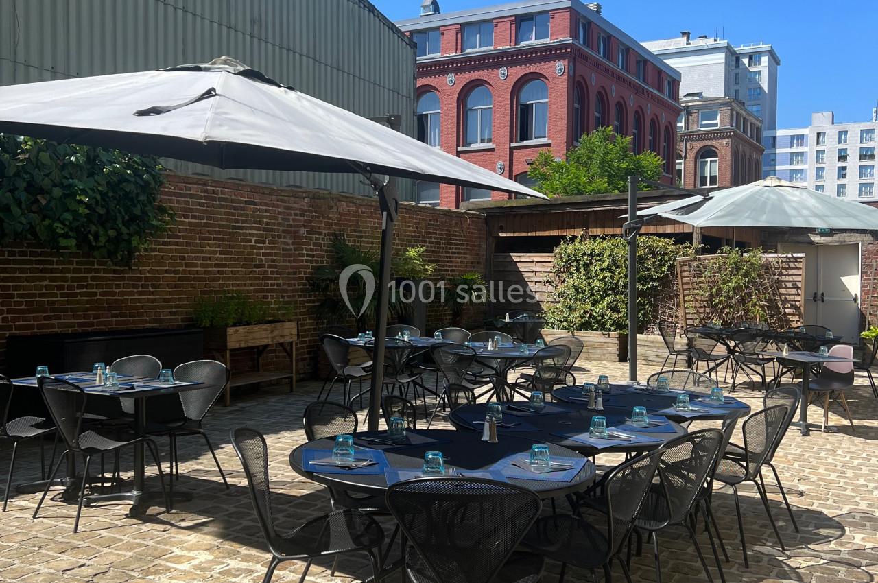 Location salle Lille (Nord) - Les Salons de l'Urban #32 Terrasse extérieure avec tables et chaises en métal, parasols ouverts, entourée de murs en briques et bâtiments urbains.