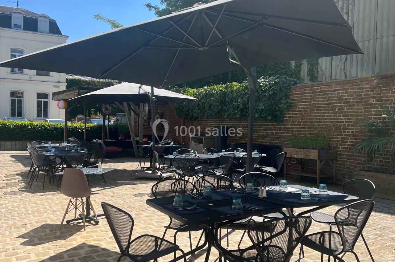 Location salle Lille (Nord) - Les Salons de l'Urban #17 Terrasse extérieure ensoleillée avec tables et chaises noires, parasols ouverts et murs en briques en arrière-plan.