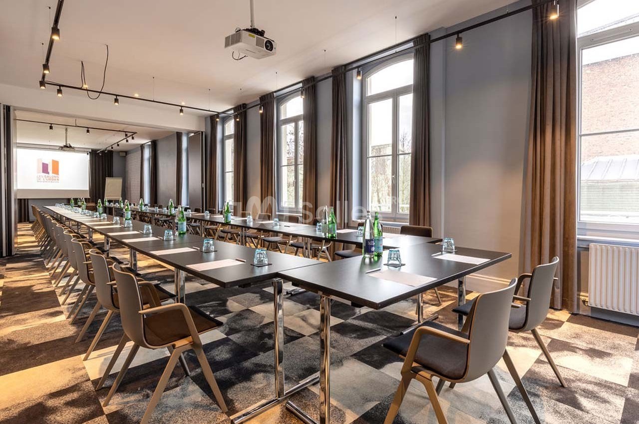 Salle de réunion lumineuse avec grandes fenêtres, tables disposées en U, chaises modernes et matériel de présentation.