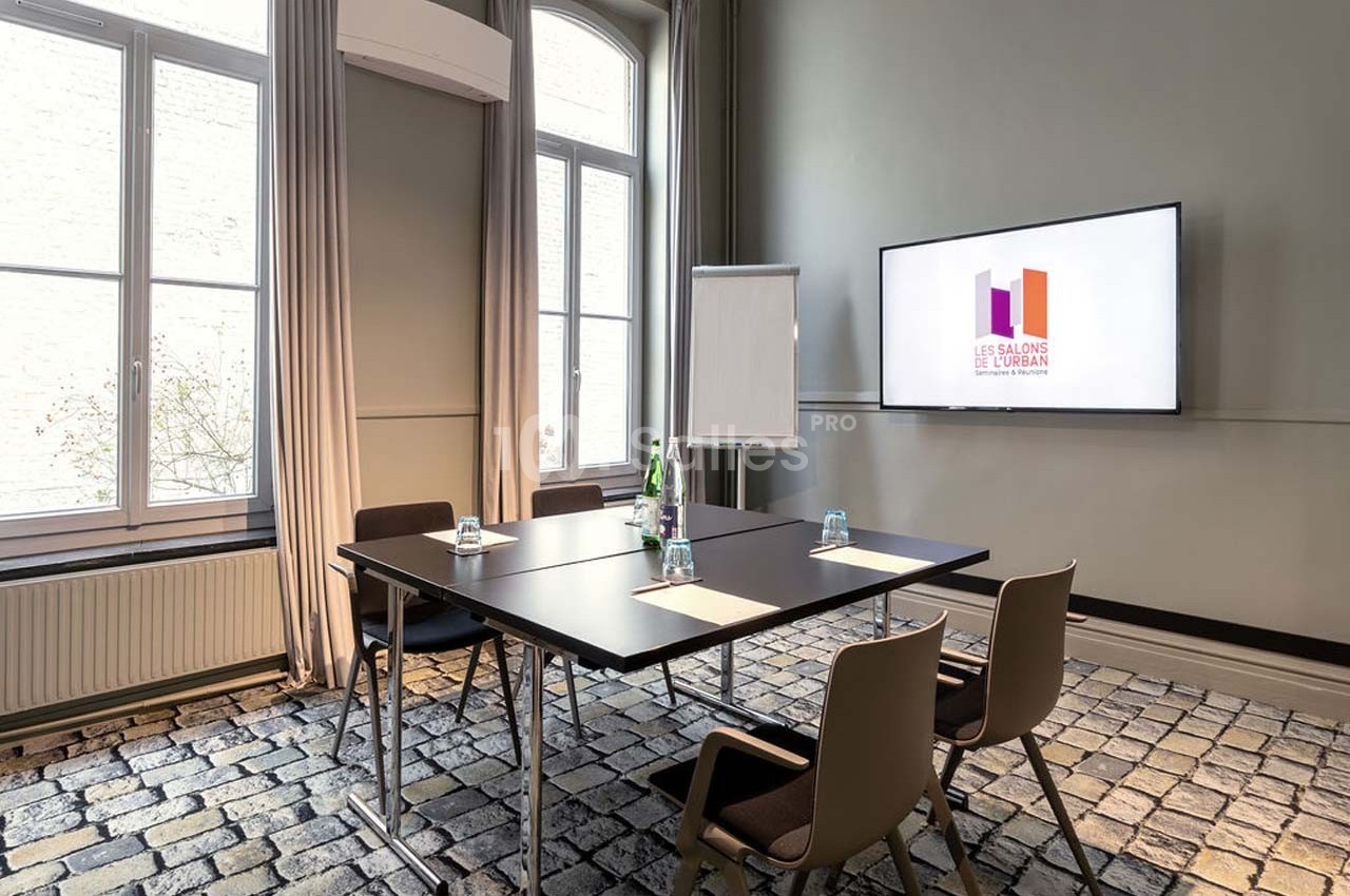 Salle de réunion lumineuse avec table, chaises, paperboard, écran affichant un logo et fenêtres donnant sur l'extérieur.