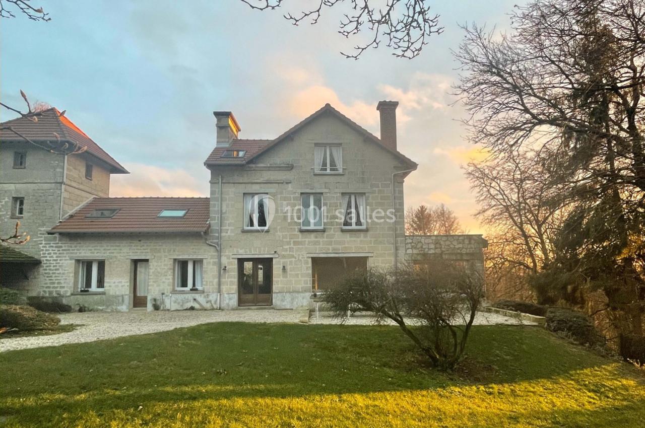 Grande maison en pierre avec toit en tuiles rouges, entourée d'un jardin verdoyant au coucher du soleil.