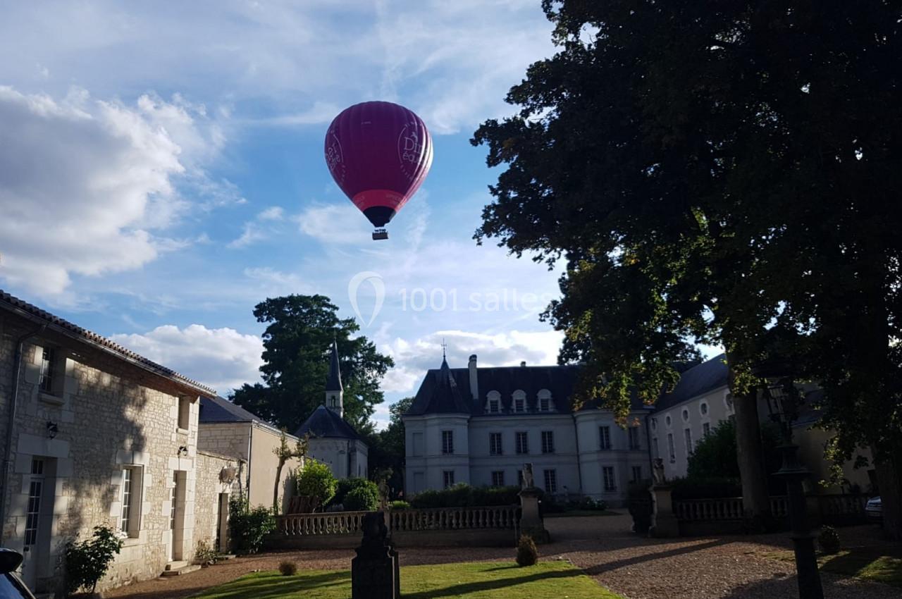 Montgolfière rouge flottant au-dessus d'une cour avec bâtiments anciens et arbres sous un ciel partiellement nuageux.