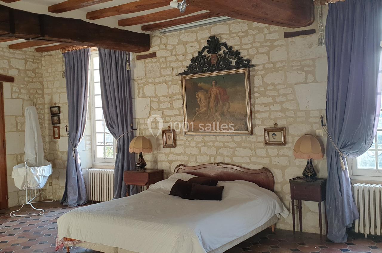 Chambre avec murs en pierre, poutres apparentes, lit double, rideaux violets et mobilier ancien.