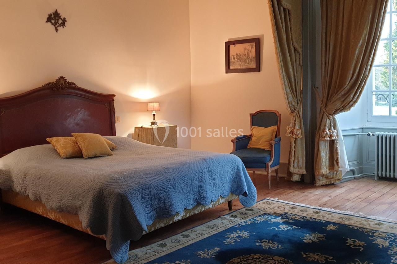 Chambre lumineuse avec lit double, fauteuil, tapis bleu, rideaux dorés et grande fenêtre donnant sur un jardin.