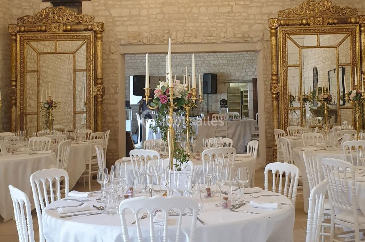 Salle de réception élégante avec tables rondes dressées, chaises blanches et grands miroirs dorés sur les murs en pierre.
