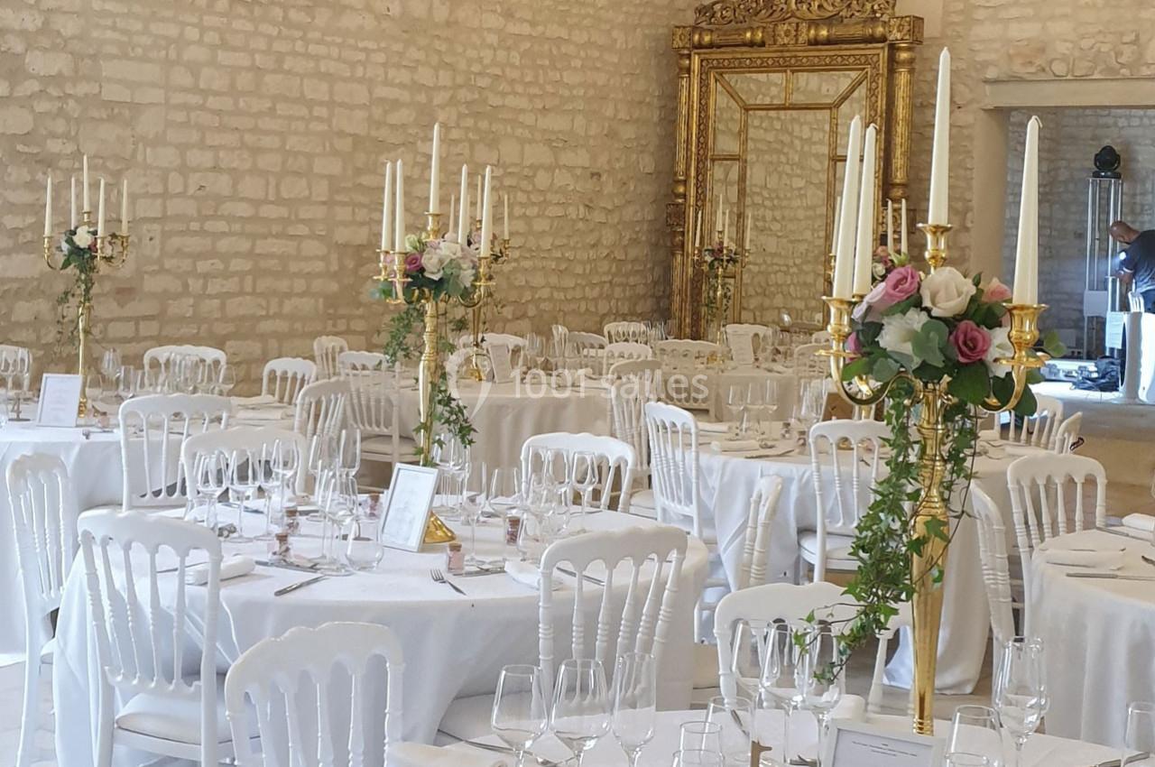 Salle de réception décorée avec des tables rondes, nappes blanches, chandeliers dorés et compositions florales.