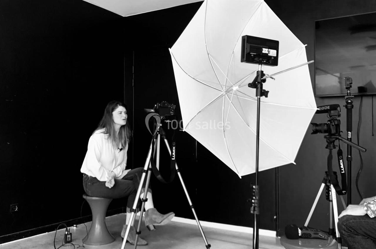 Une femme assise devant un appareil photo sur trépied, éclairée par un parapluie lumineux dans un studio sombre.