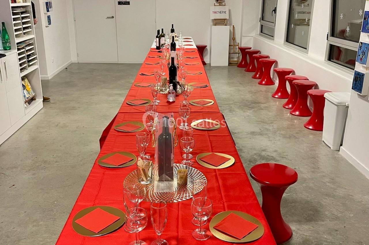 Grande table rectangulaire décorée pour un repas festif, avec nappes rouges, verres et assiettes dorées dans une salle…