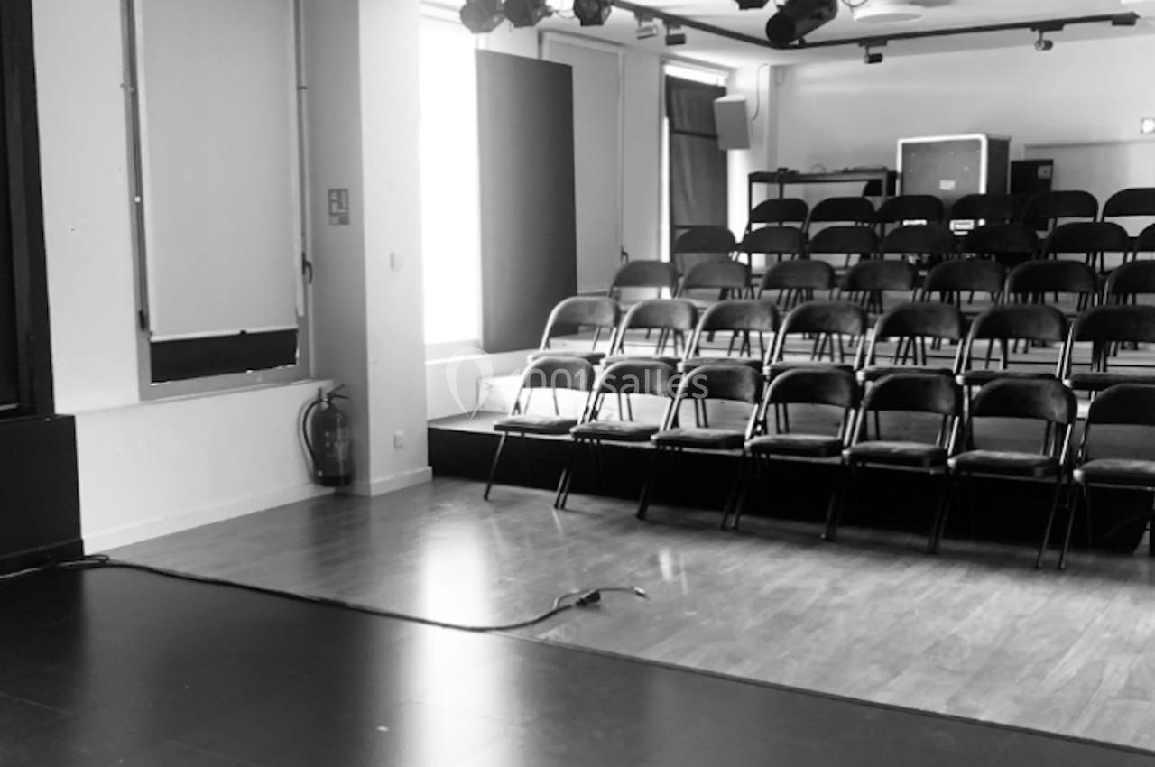 Salle de spectacle vide avec des rangées de chaises noires face à une scène, éclairage suspendu au plafond.