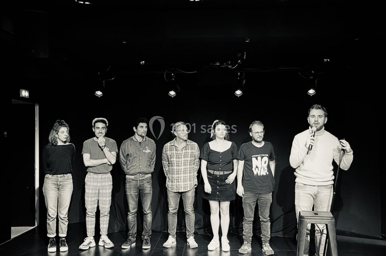 Un groupe de sept personnes debout sur une scène sombre, un homme tenant un micro à droite.