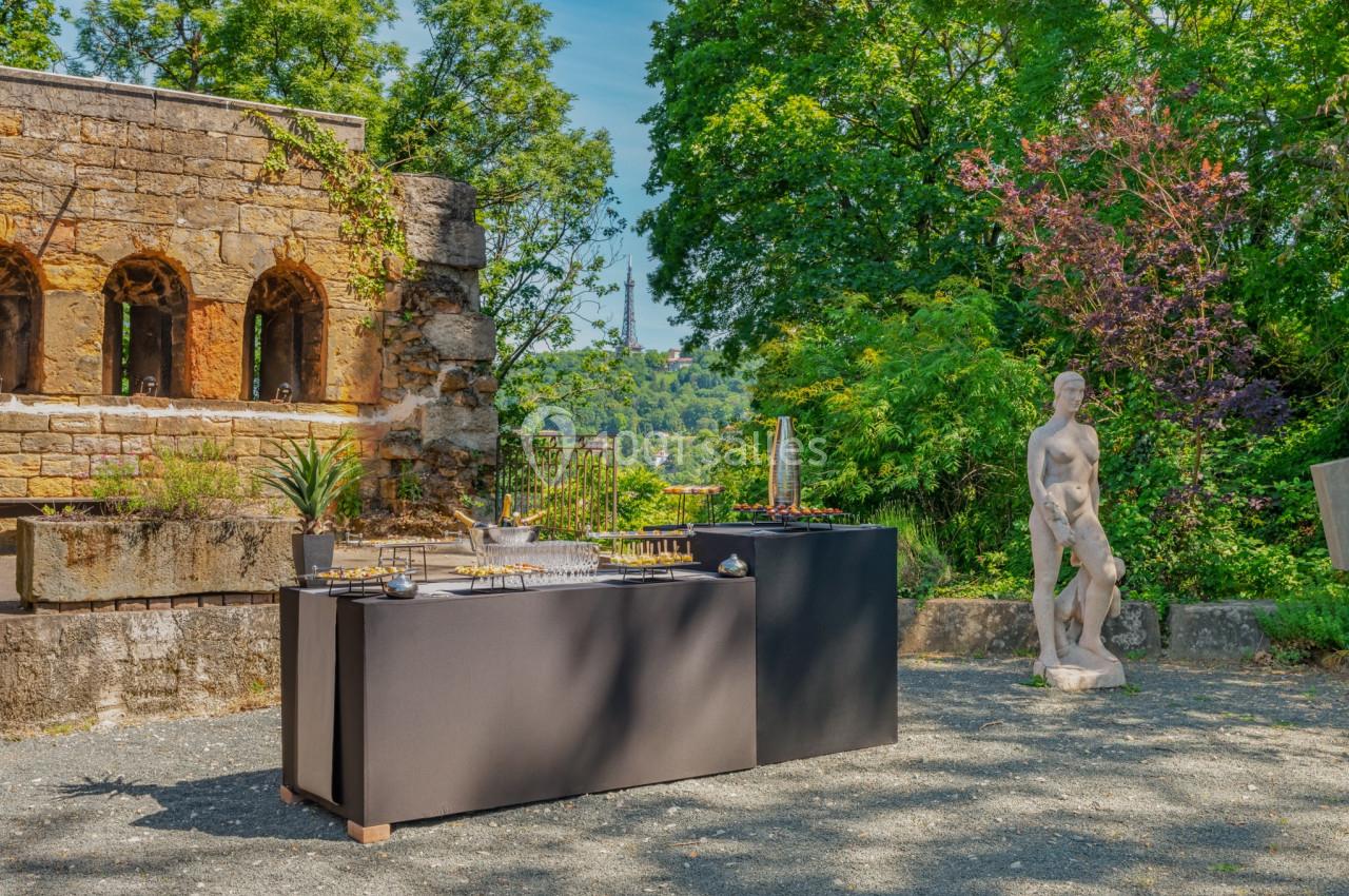 Deux tables noires avec des objets exposés dans un jardin, près d'une statue et d'un mur en pierre ancienne.