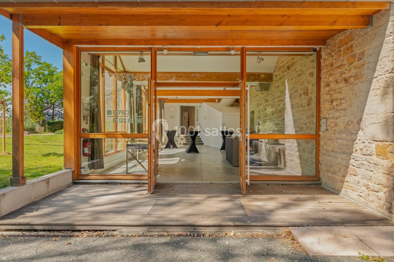 Entrée vitrée d'un bâtiment en pierre, donnant sur une salle avec des tables hautes et un espace extérieur verdoyant.