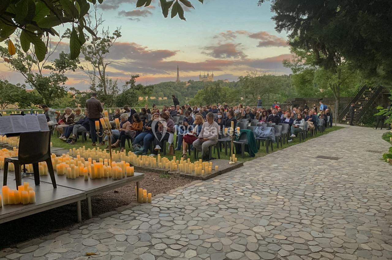 Un public assis en plein air au coucher du soleil, entouré de bougies, face à une scène avec des partitions.