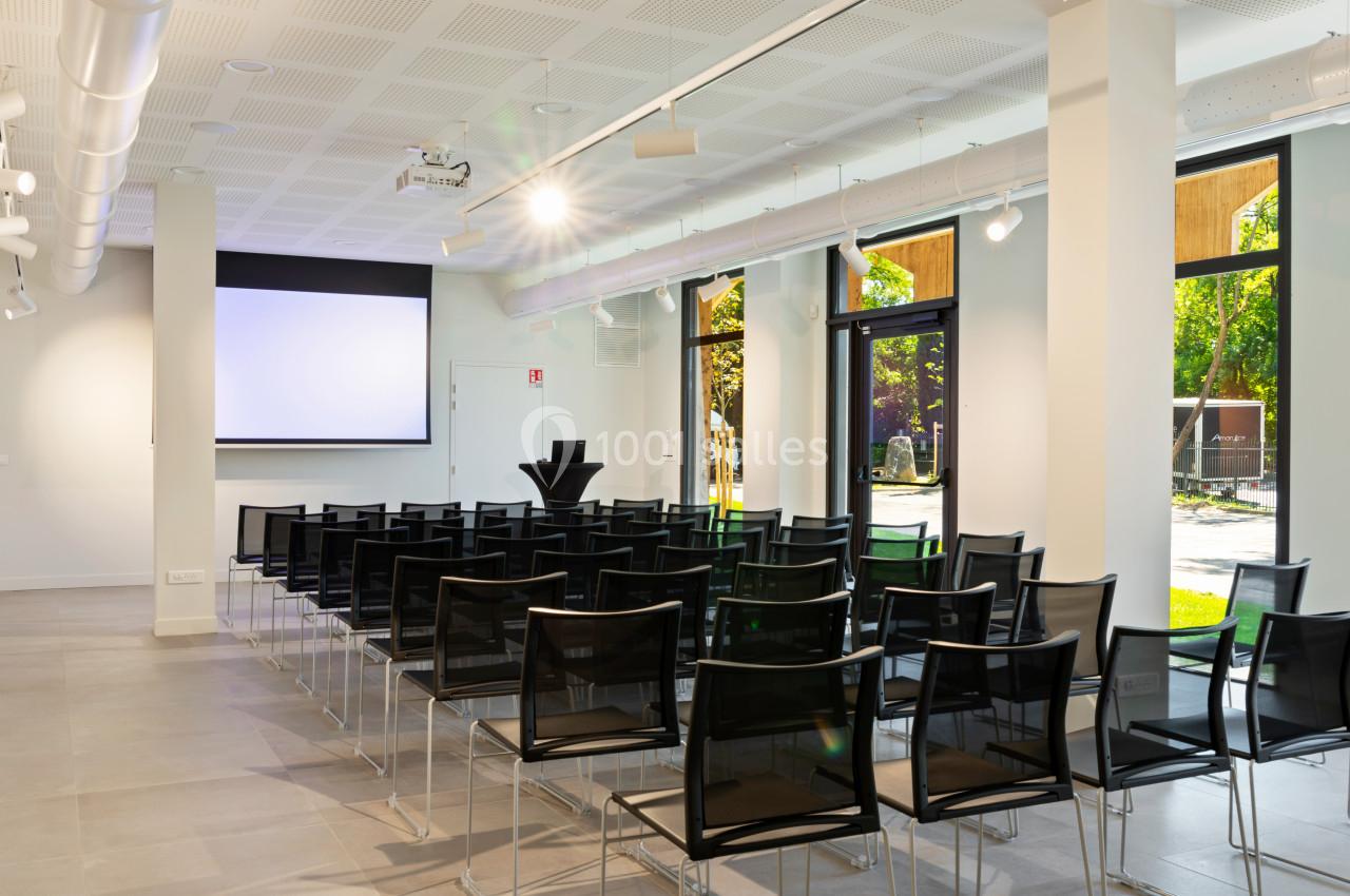 Salle de conférence lumineuse avec des chaises noires alignées, un écran de projection et de grandes fenêtres donnant sur l…