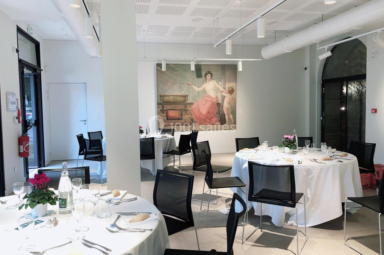 Salle lumineuse aménagée pour un repas, avec des tables rondes dressées et une grande peinture murale en arrière-plan.