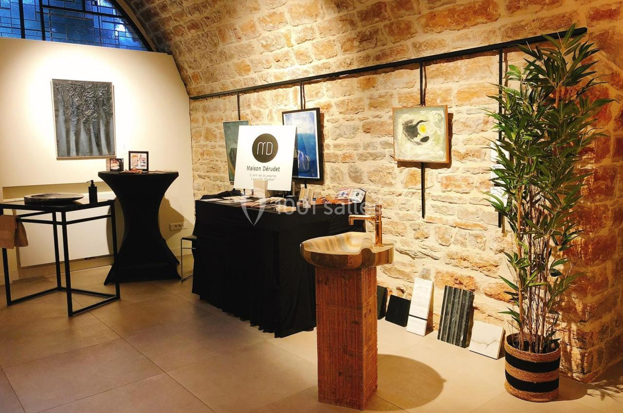 Espace d'exposition avec des œuvres d'art accrochées sur un mur en pierre, une table et un présentoir.