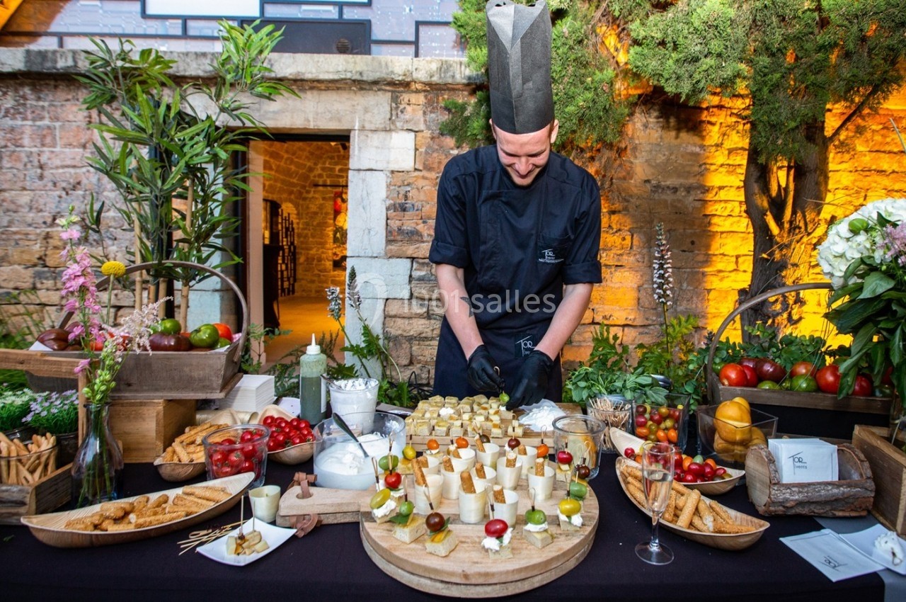 Location salle Lyon 9 (Rhône) - Le Fort de Vaise #5 Un chef en tenue noire prépare un buffet de bouchées apéritives sur une table décorée en extérieur.