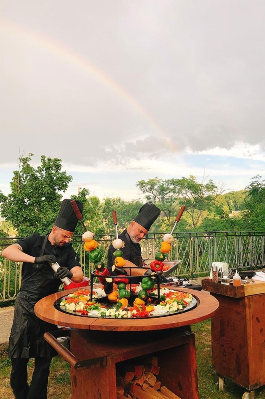 Location salle Lyon 9 (Rhône) - Le Fort de Vaise #6 Deux chefs cuisinent des brochettes de légumes et de viande sur un barbecue circulaire en plein air sous un arc-en-ciel.