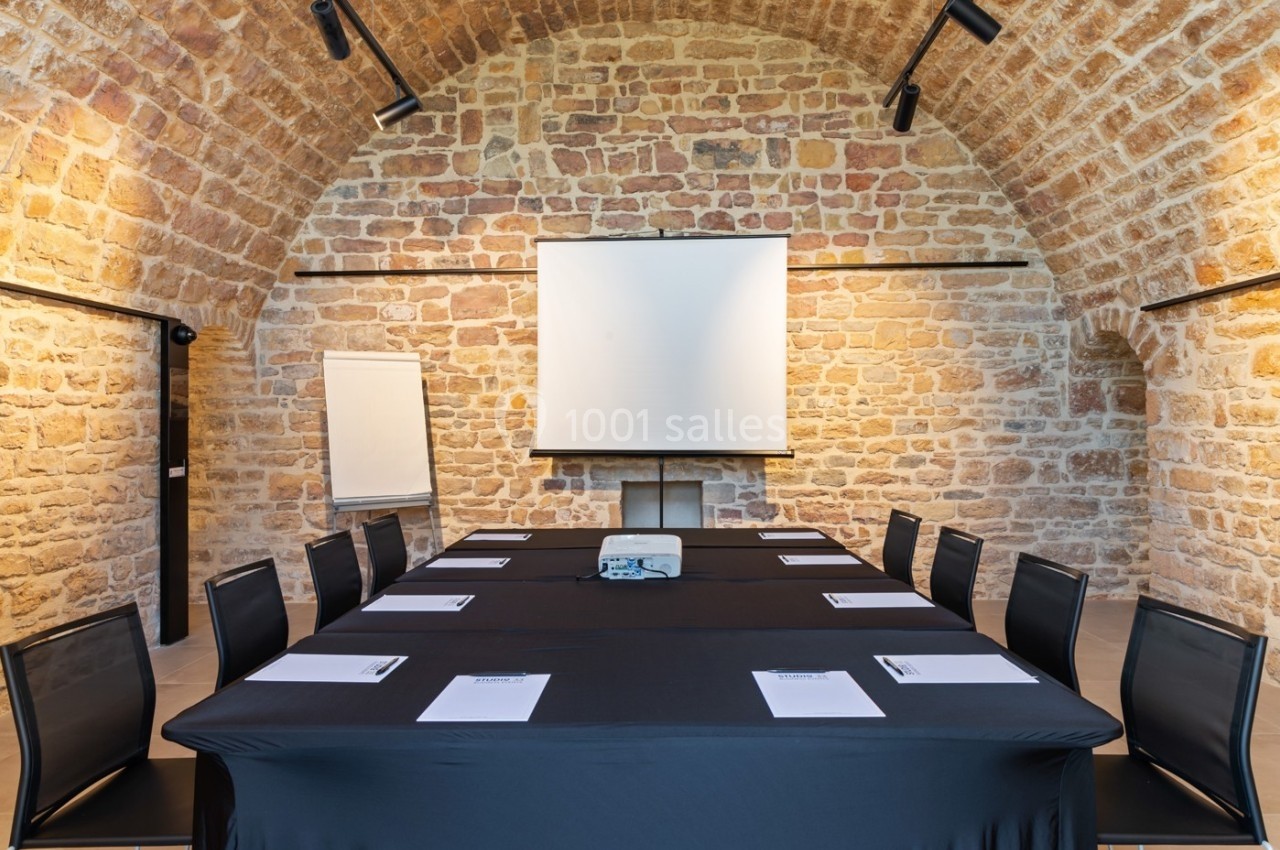 Location salle Lyon 9 (Rhône) - Le Fort de Vaise #11 Salle de réunion avec table rectangulaire, chaises noires, projecteur, écran blanc et murs en pierre apparente.