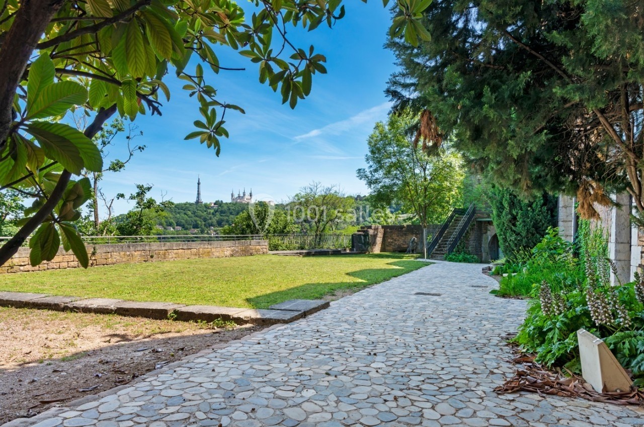 Location salle Lyon 9 (Rhône) - Le Fort de Vaise #12 Allée pavée bordée de verdure menant à un espace ouvert avec vue sur une colline et des bâtiments au loin.