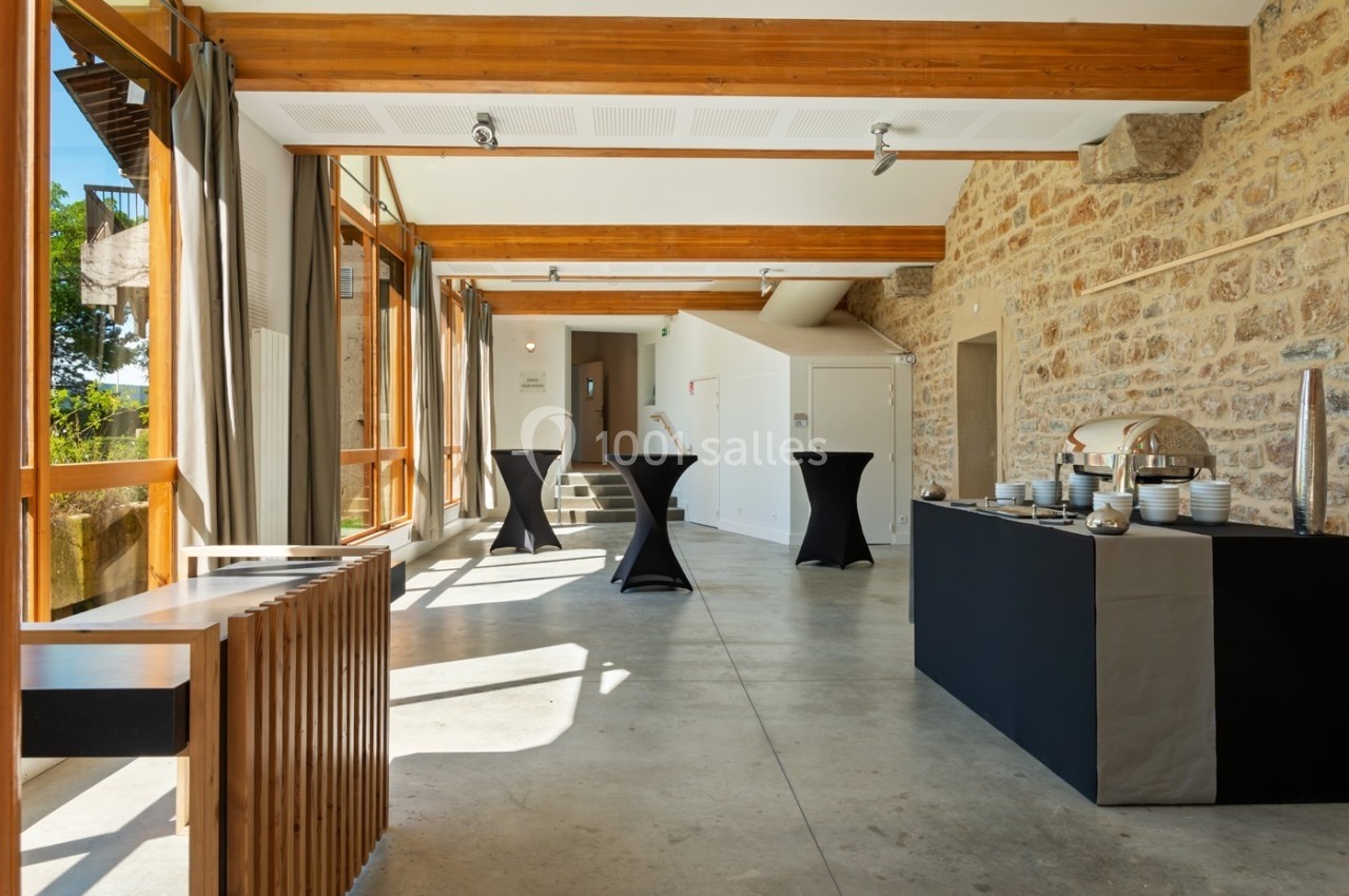 Location salle Lyon 9 (Rhône) - Le Fort de Vaise #15 Salle lumineuse avec murs en pierre, grandes baies vitrées, tables hautes noires et buffet dressé sur la droite.