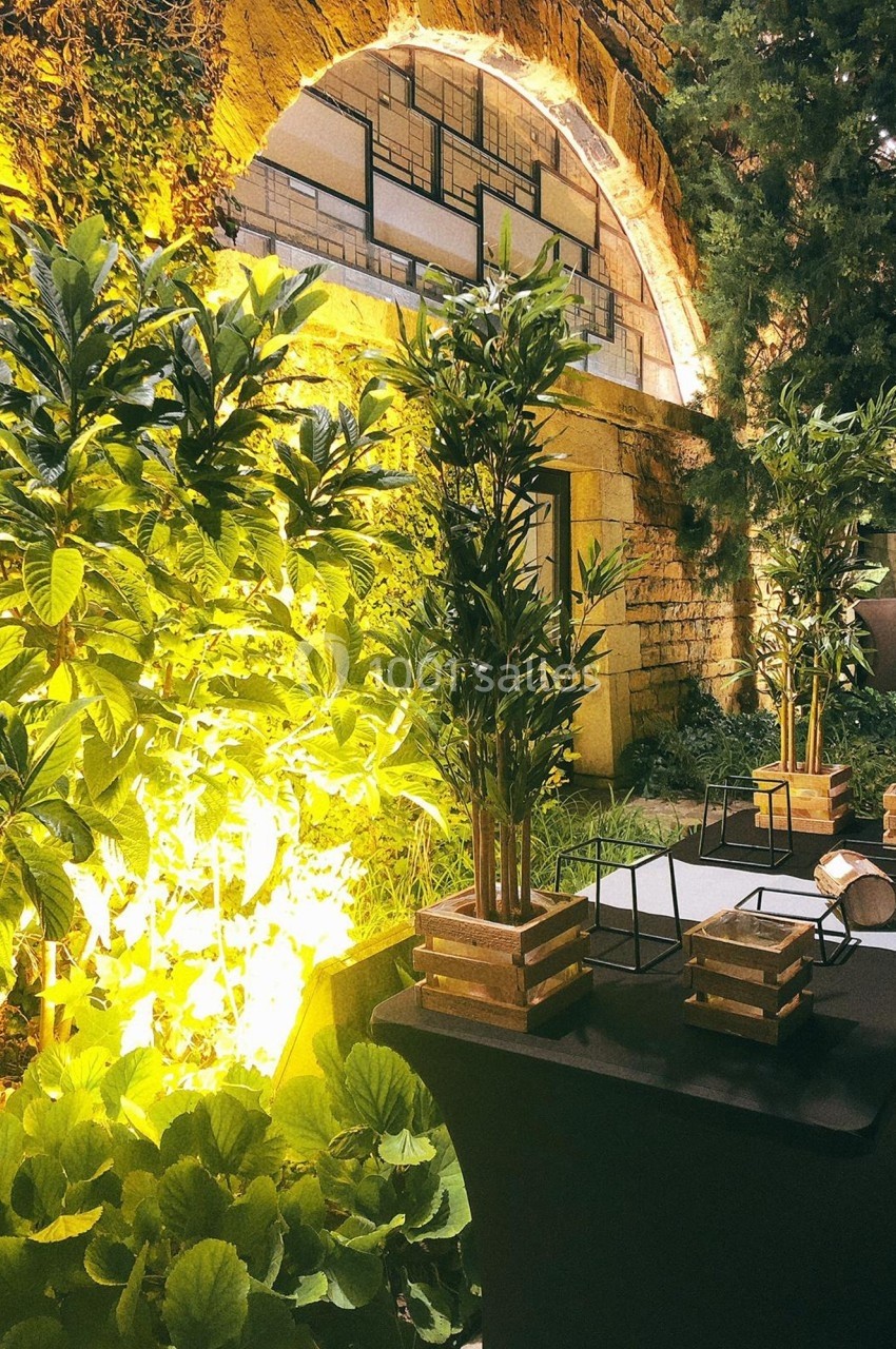 Location salle Lyon 9 (Rhône) - Le Fort de Vaise #24 Espace extérieur aménagé avec des plantes, des tables noires et un éclairage chaleureux près d'une arche en pierre.