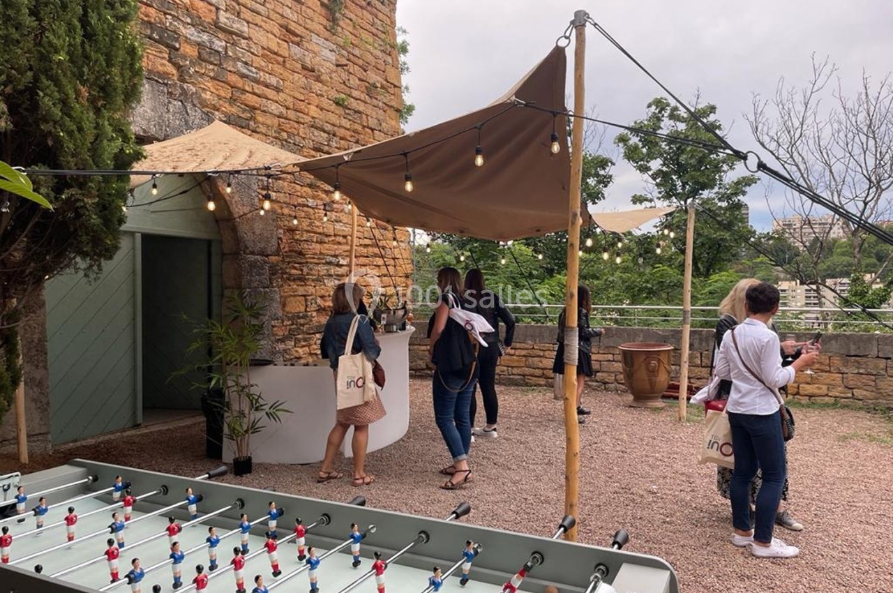 Location salle Lyon 9 (Rhône) - Le Fort de Vaise #26 Des personnes discutent près d'un stand en extérieur, sous une toile tendue, avec un baby-foot au premier plan.