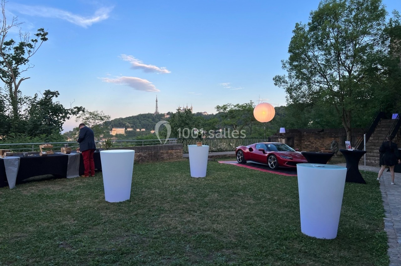 Location salle Lyon 9 (Rhône) - Le Fort de Vaise #27 Vue d'un jardin avec une voiture sportive rouge, des tables hautes et un paysage de colline en arrière-plan au crépuscule.