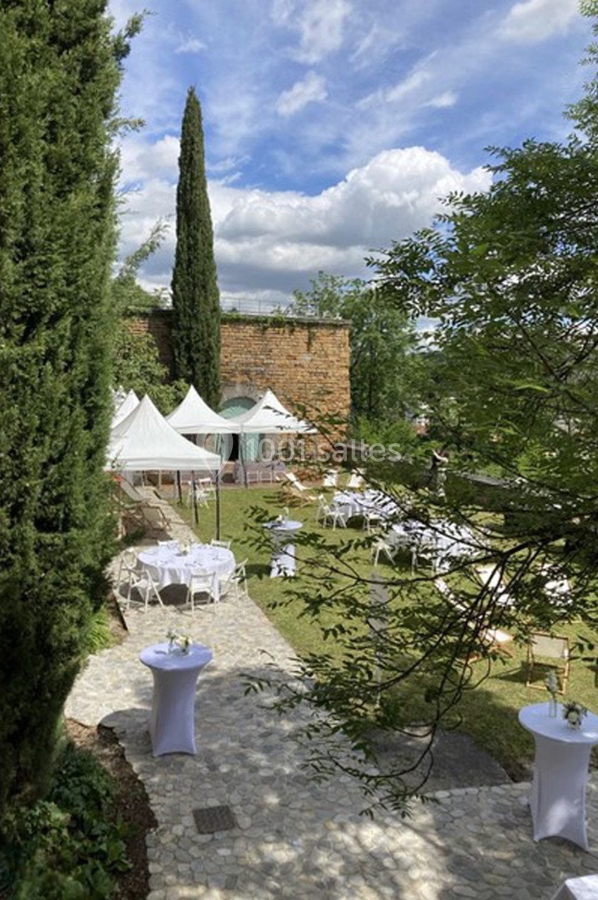 Location salle Lyon 9 (Rhône) - Le Fort de Vaise #33 Jardin aménagé avec des tables et des tentes blanches, entouré de verdure et d'un mur en pierre sous un ciel partiellement…