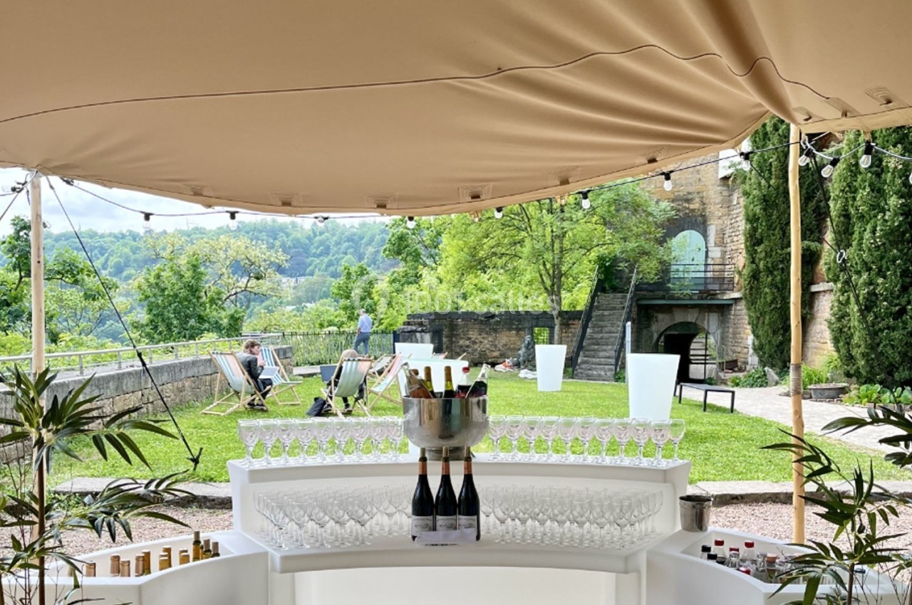 Location salle Lyon 9 (Rhône) - Le Fort de Vaise #34 Bar extérieur sous une tente beige avec des verres et des bouteilles, vue sur un jardin verdoyant en arrière-plan.