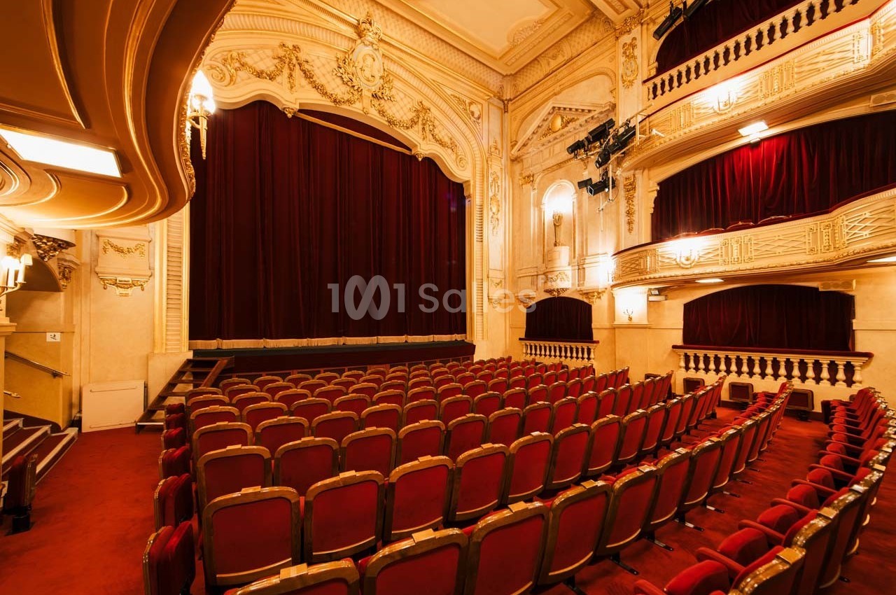 Salle de théâtre avec rangées de fauteuils rouges, scène avec rideau fermé et décorations intérieures élégantes.