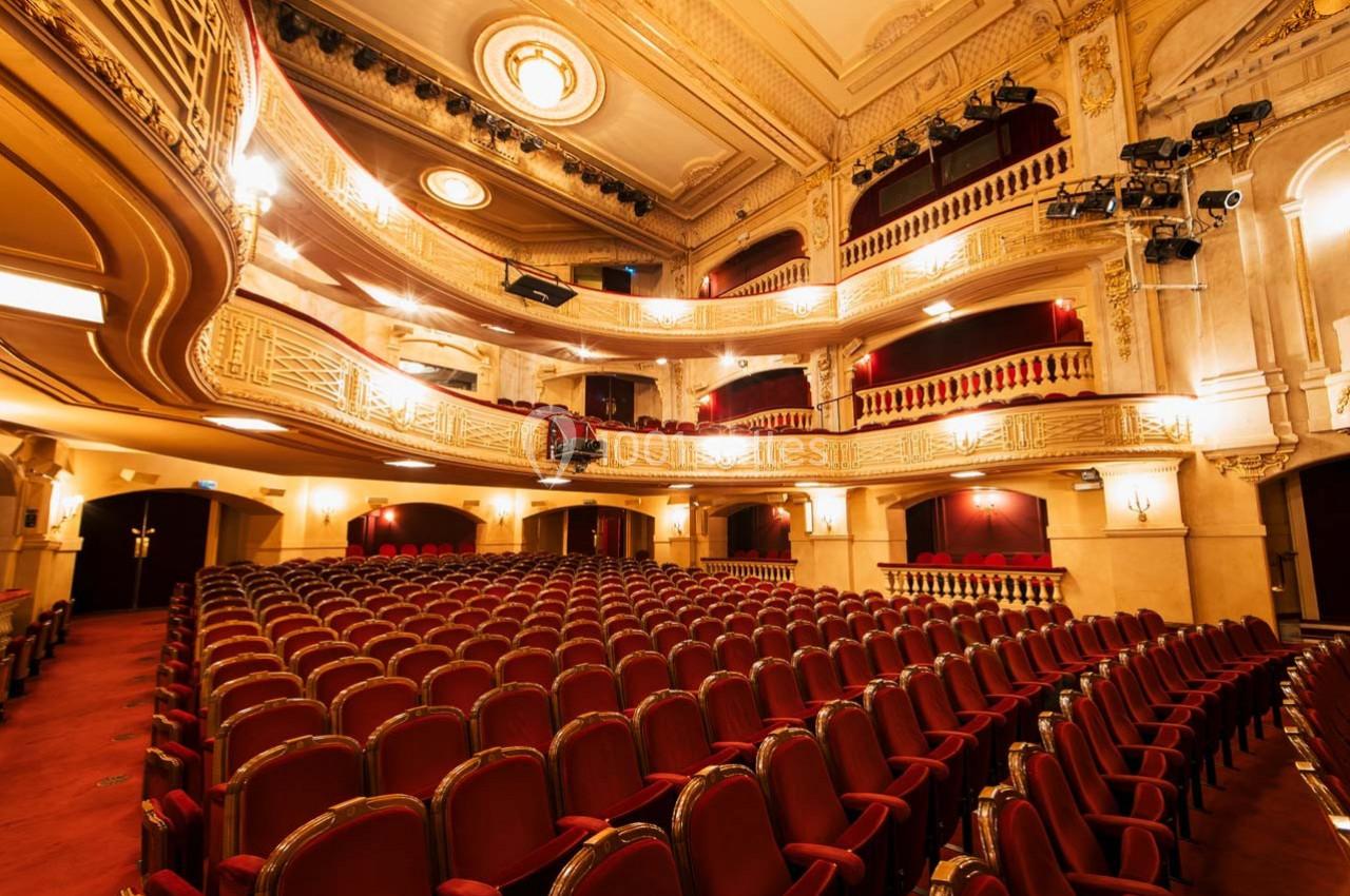 Salle de théâtre vide avec rangées de fauteuils rouges, balcons ornés et éclairage chaleureux.