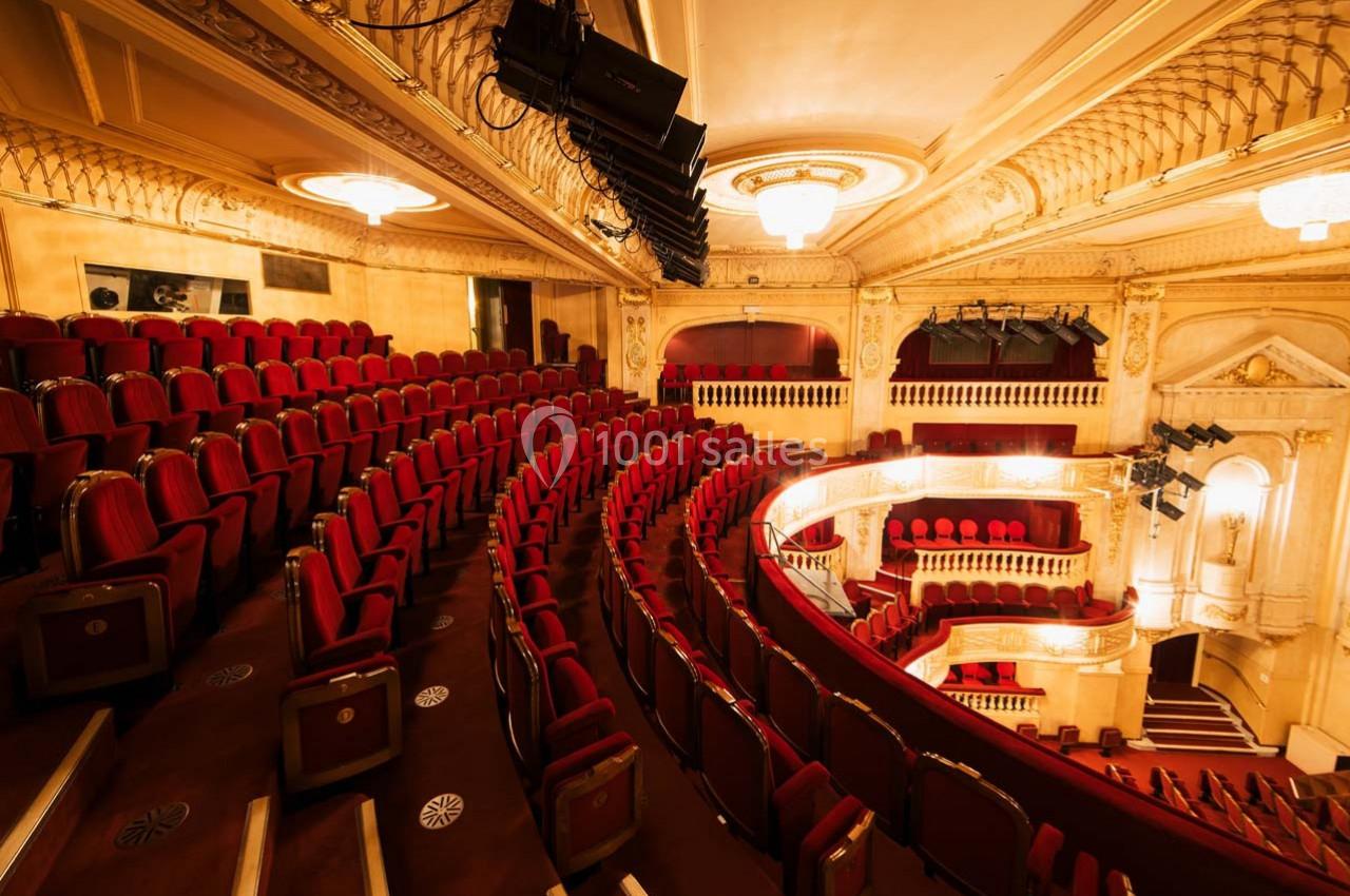 Salle de théâtre avec des rangées de sièges rouges, balcons décorés et éclairage chaleureux.