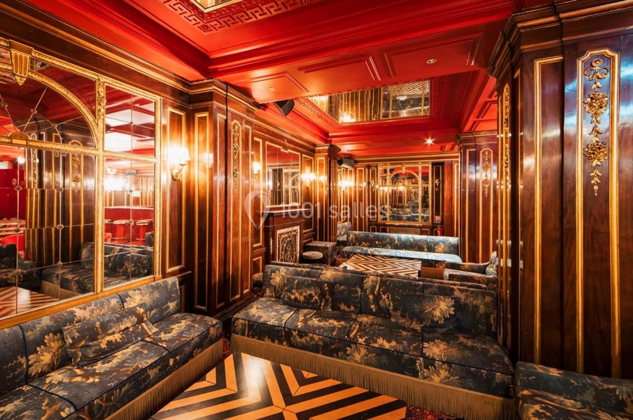 Salon élégant avec boiseries, miroirs, canapés à motifs floraux et éclairage chaleureux dans un décor rouge et doré.